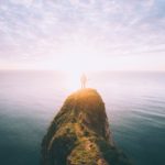 Licht am Horizont: Person steht auf einem Felsen über dem Meer, im Hintergrund geht die Sonne über dem Meer auf