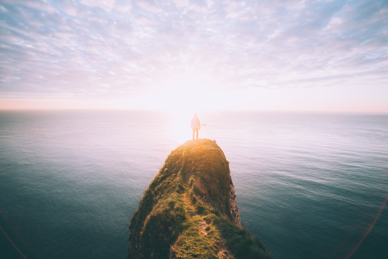 Licht am Horizont: Person steht auf einem Felsen über dem Meer, im Hintergrund geht die Sonne über dem Meer auf